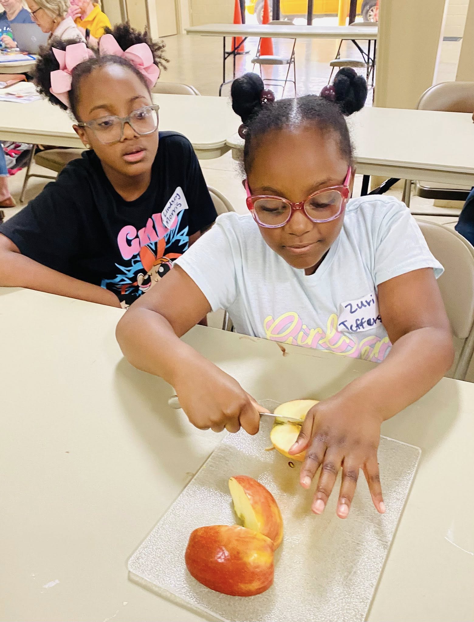 A girl watching another girl cut an apple.
